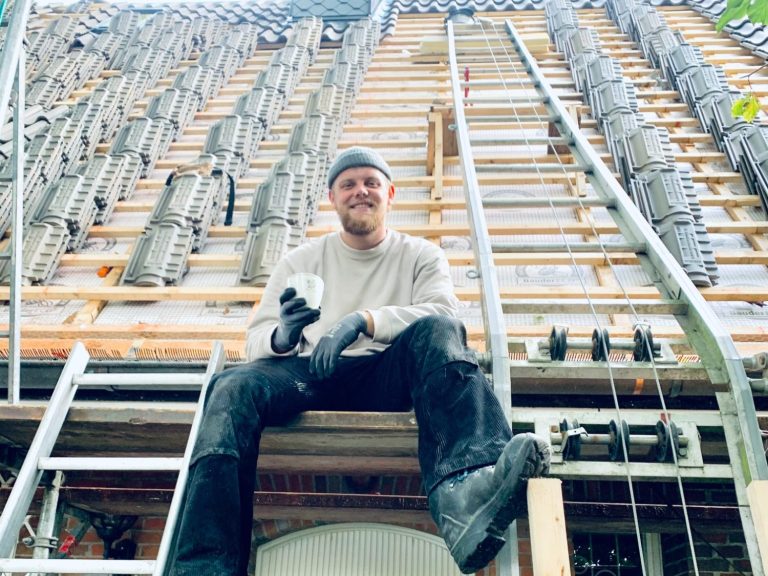 Handwerker am Umdecken eines Dachs Handwerker sitzt auf einer Leiter vor einem Gerüst und hält ein Werkzeug in der Hand.