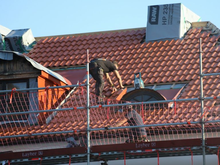 Dachsanierung Dachdecker arbeitet auf einem Gerüst und deckt ein Dach mit roten Ziegeln.